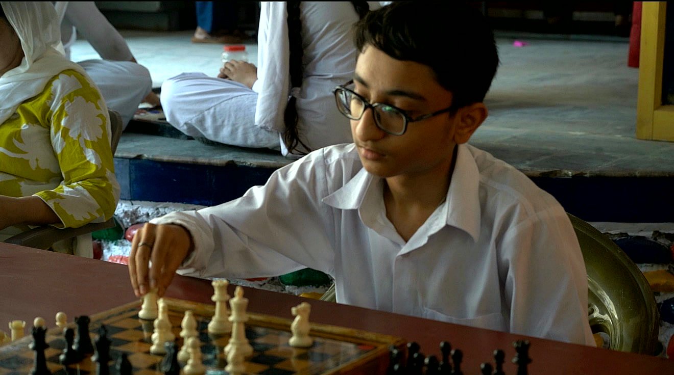 A senior student in white uniform reaching to make a move at a chess board, other students playing on the floor behind him