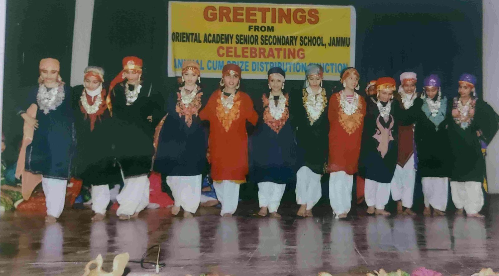 Children in Dogri folk costume lined up on stage under a school banner
