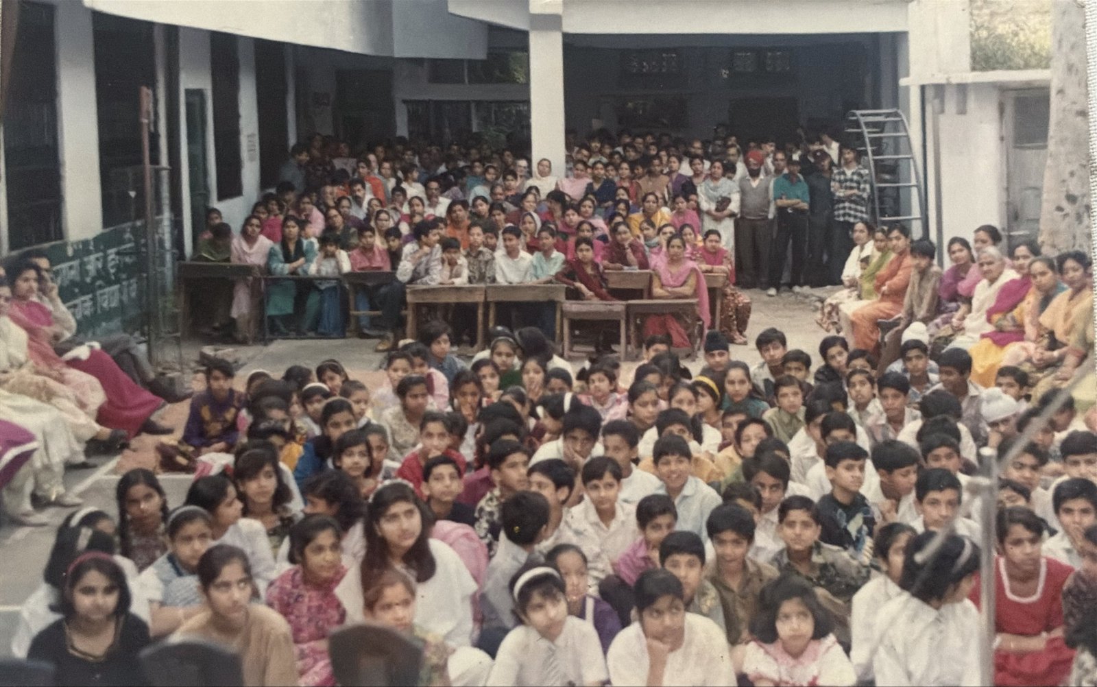 The whole school gathered in the courtyard for a function in the 1980s: children seated on the ground, parents on benches behind, the old building and its verandah around them