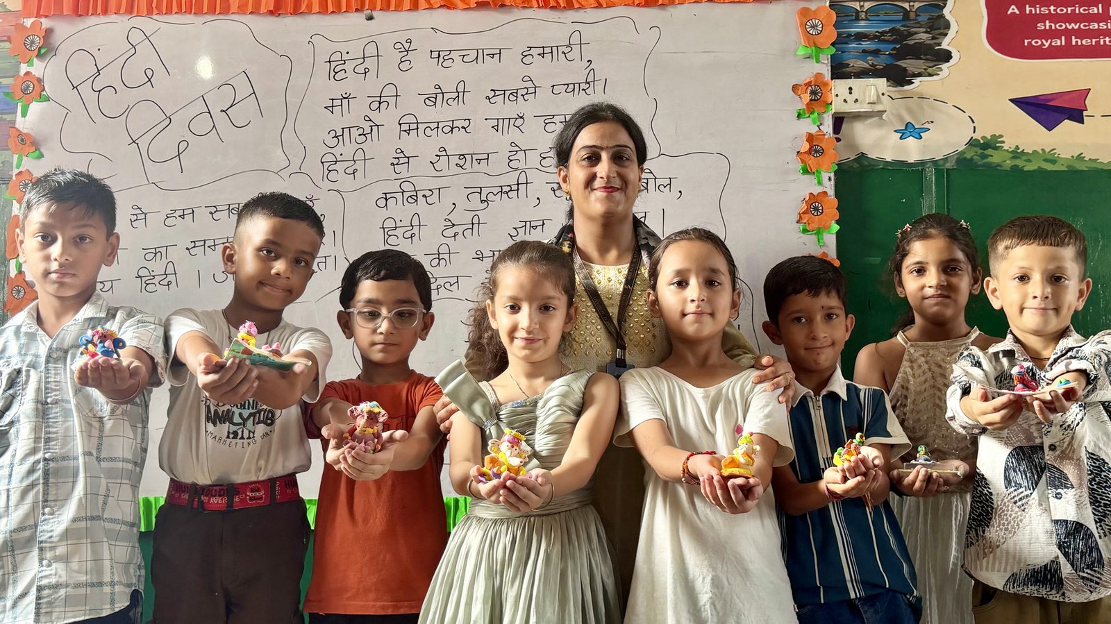 A teacher and eight children in a classroom, each holding out on open palms a small clay Ganesha they made, in front of a hand-lettered board reading Hindi Diwas with a Hindi poem