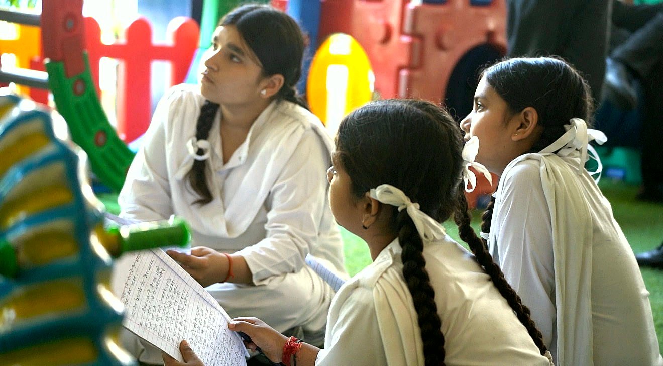 Three girls in white uniform sitting on the grass in the play area, looking up and listening, one of them holding a handwritten page