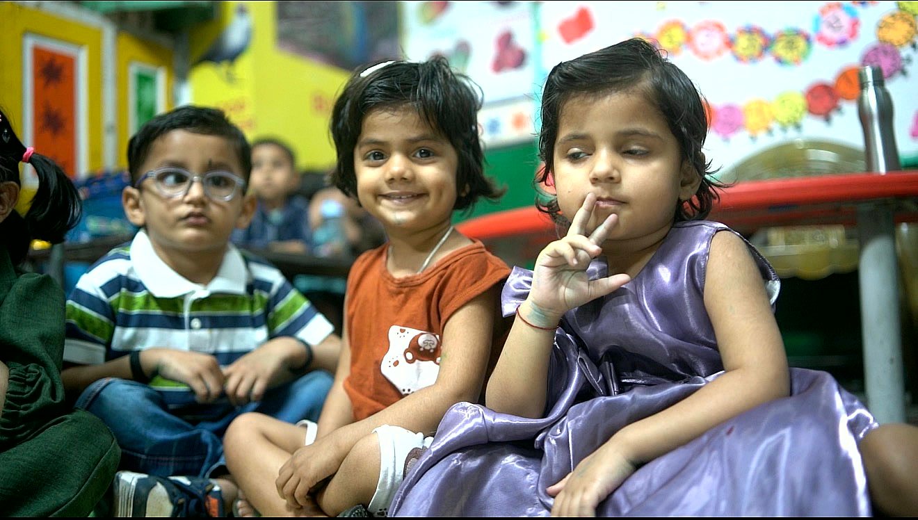 Three nursery children sitting together on the floor, one of them smiling straight at the camera, cut-paper decorations on the wall behind them