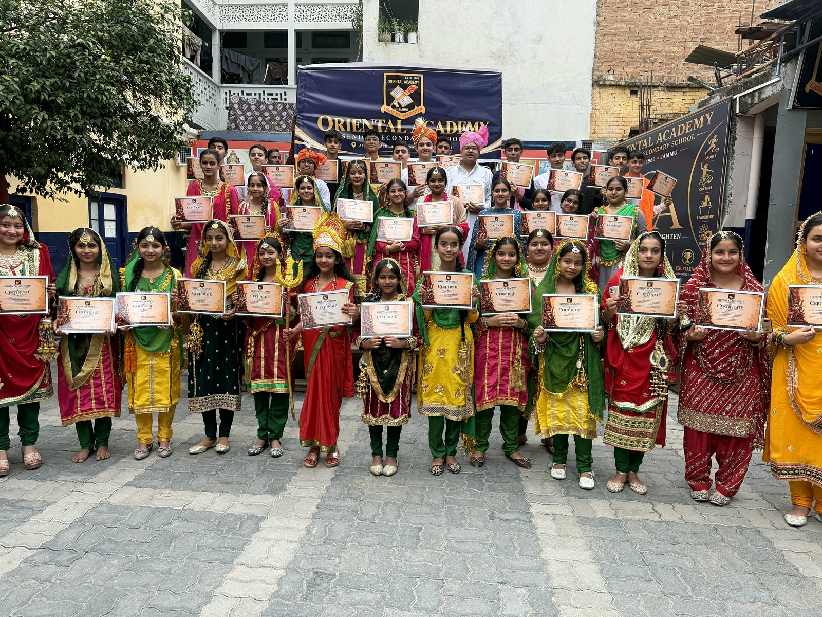 A large group of Oriental Academy students in traditional Dogra dress holding their certificates in front of the school banner