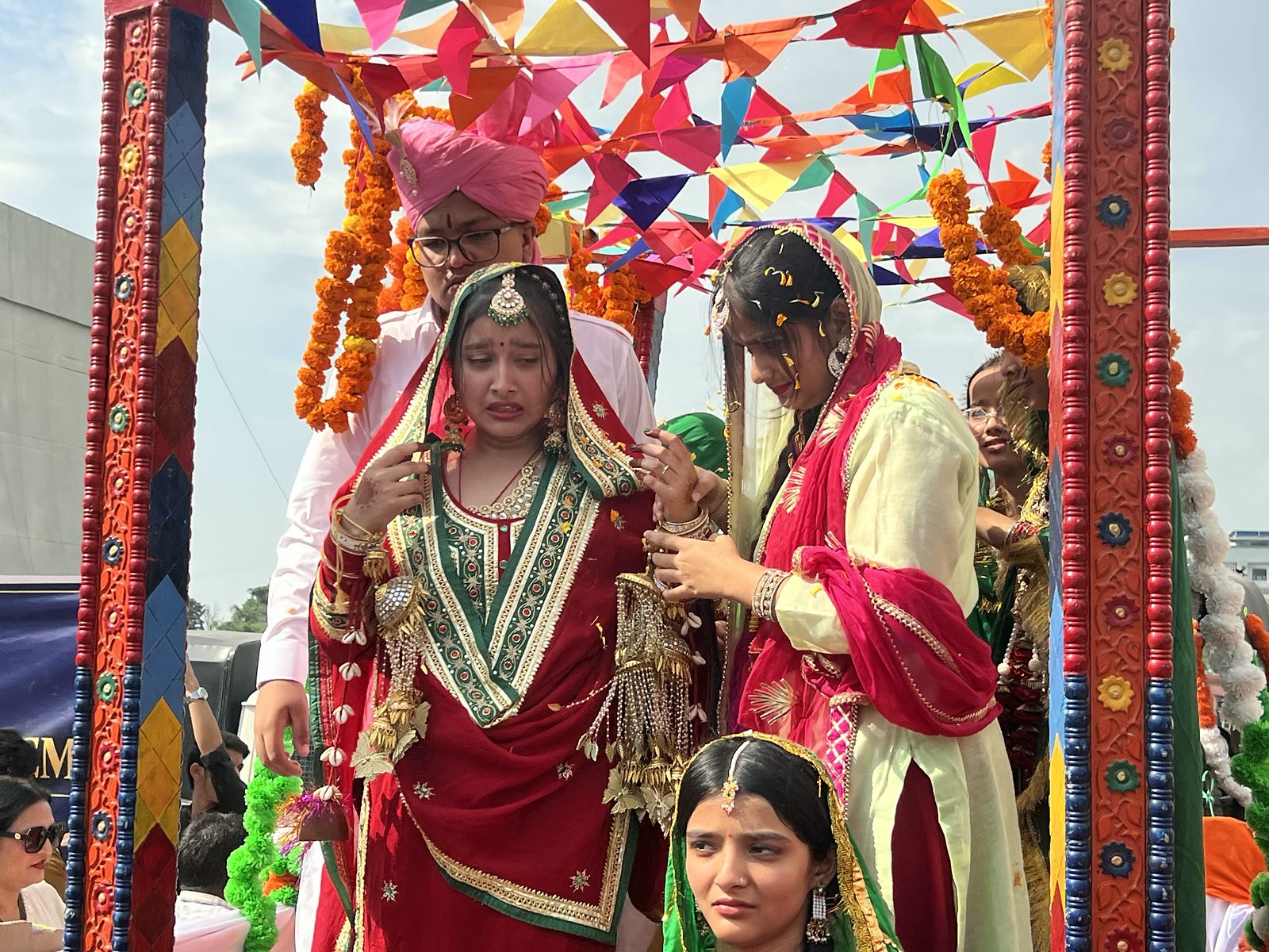 A student dressed as a Dogra bride in red, seated in a decorated doli with the wedding party around her