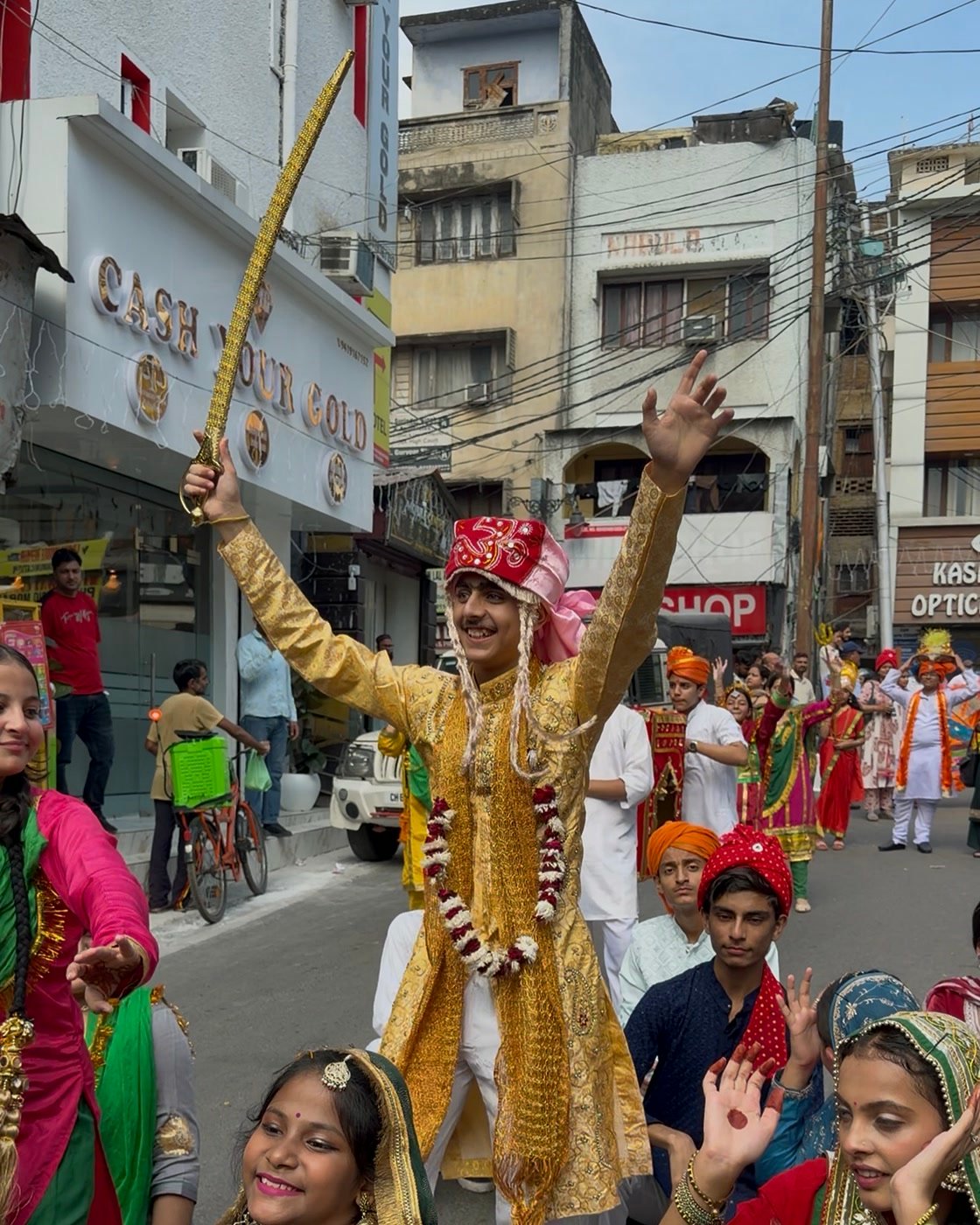The student playing the groom, in a golden sherwani and red turban with a garland, raising a sword as he dances with girls in Dogra dress