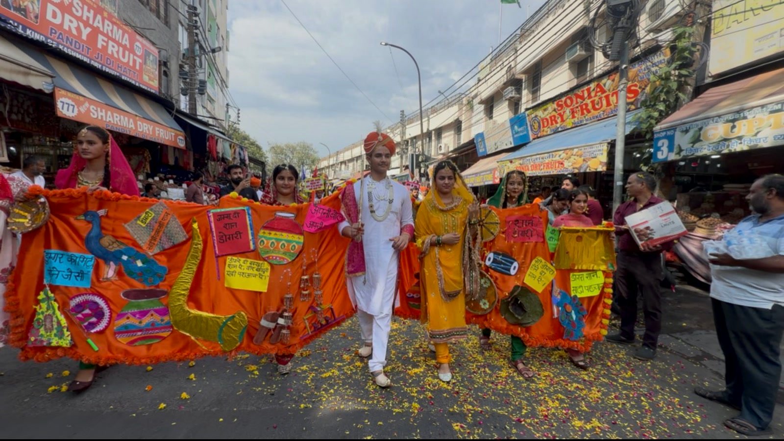 The student king and the girls holding his long orange robe walking through a Jammu market, with flower petals on the road