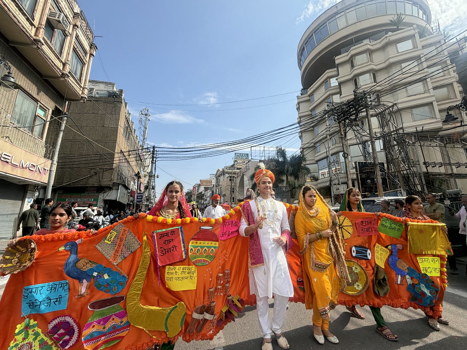 A student dressed as a king in white with a saffron turban, walking through a Jammu market with a long orange robe of handmade Dogra symbols and Dogri placards held out on both sides by students in traditional dress
