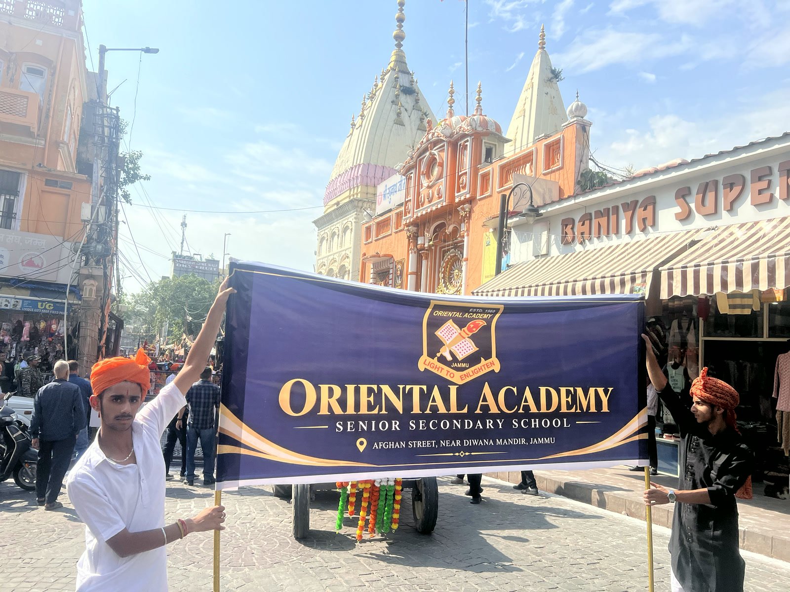 Oriental Academy students holding the school banner in a Jammu market street, with a temple behind them