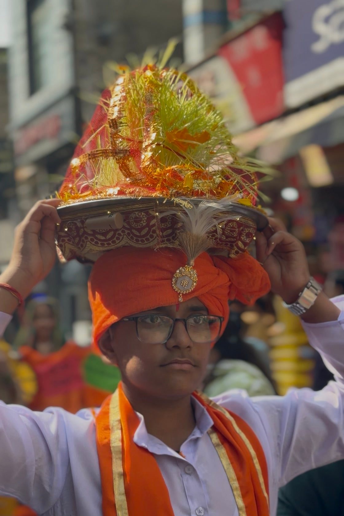 A student in an orange turban and white kurta carrying a decorated thaal on his head