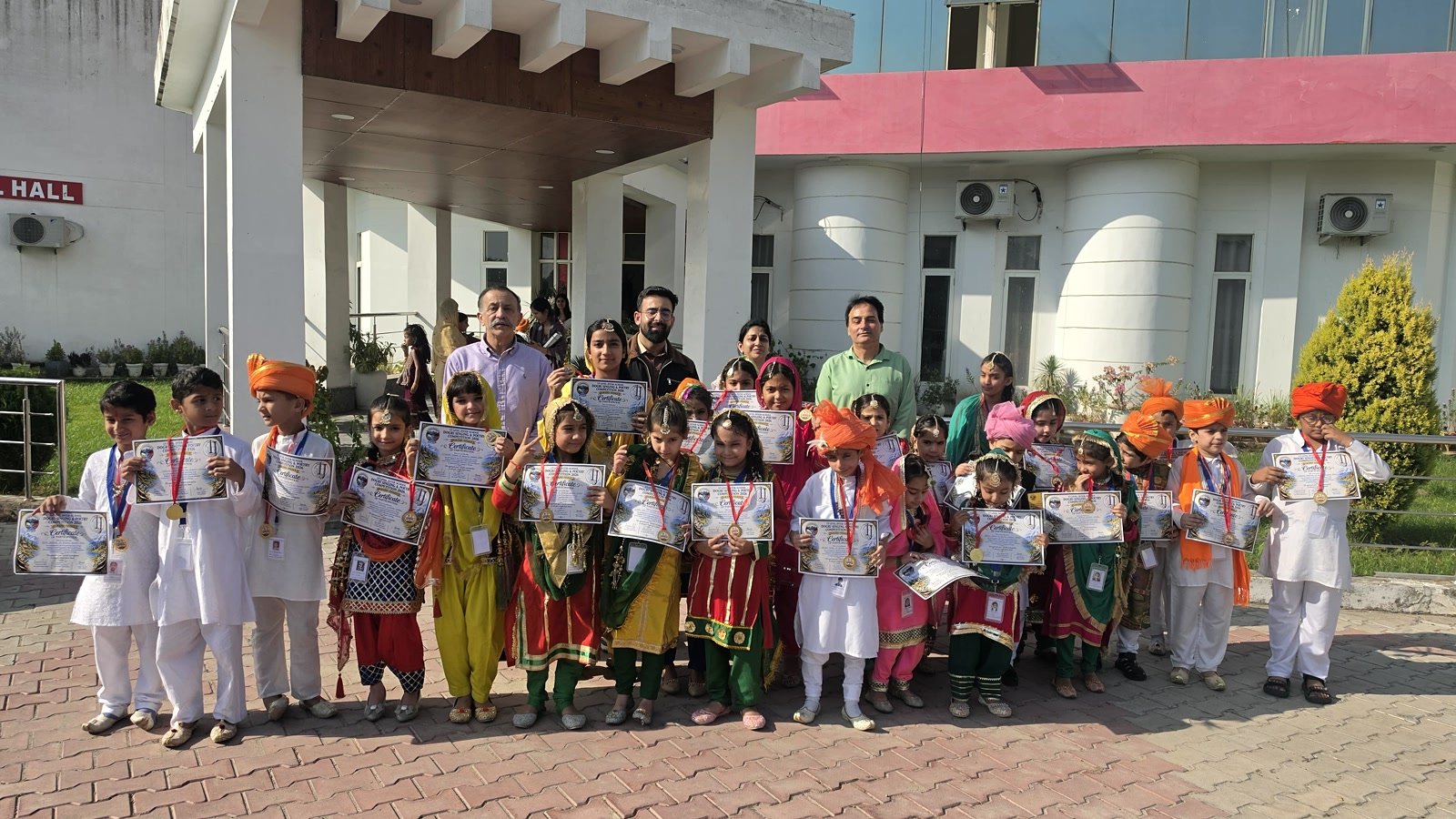 A large group of Oriental Academy students in traditional dress holding certificates outside the K.L. Saigal Hall