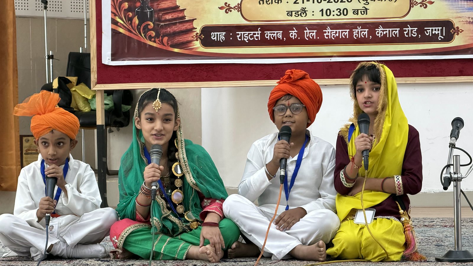 Four young students in Dogra dress seated at microphones on stage