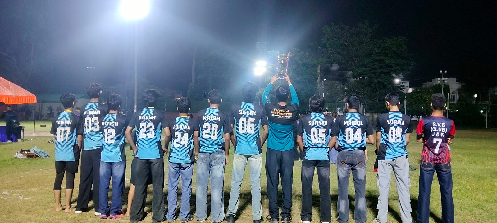 Players seen from behind, their names on their jerseys, with the runners-up trophy held high under the floodlights