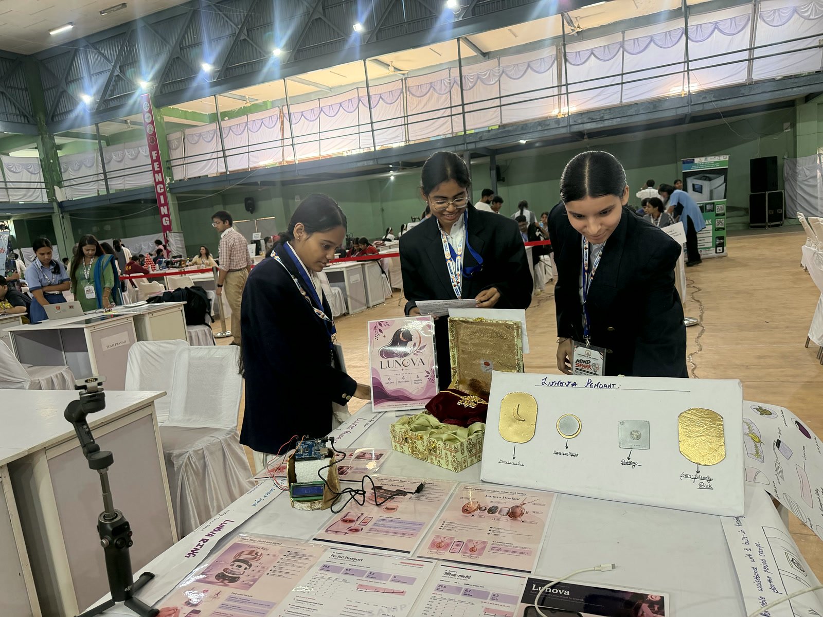 Three Oriental Academy students at the Lunova stall, beside a display card of the jewellery designs