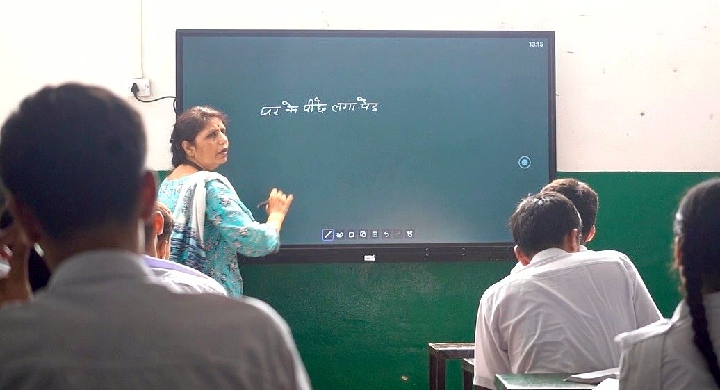 A teacher writing on a large digital display board at the front of a senior class, the line she is teaching on the screen, students following from their desks