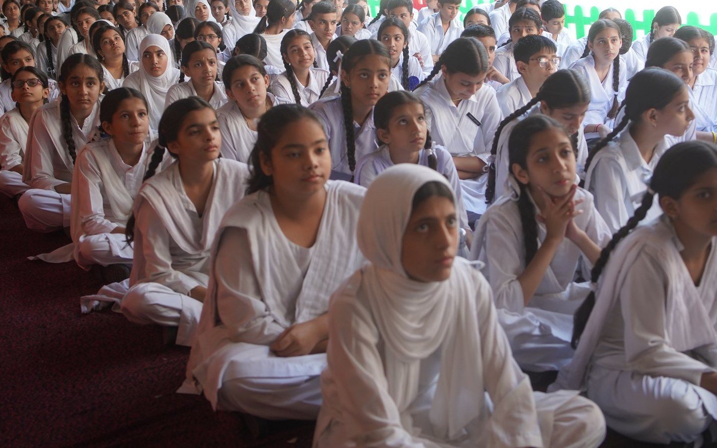 Students sitting cross-legged on the floor in the covered courtyard at assembly, listening towards the stage