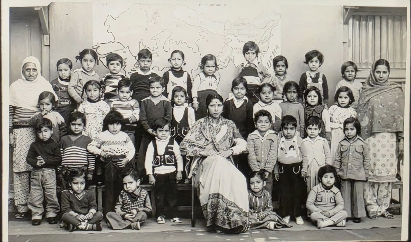 A junior class of 1982 in black and white: rows of young children in sweaters around their teacher, seated at the centre, with two helpers standing at either end