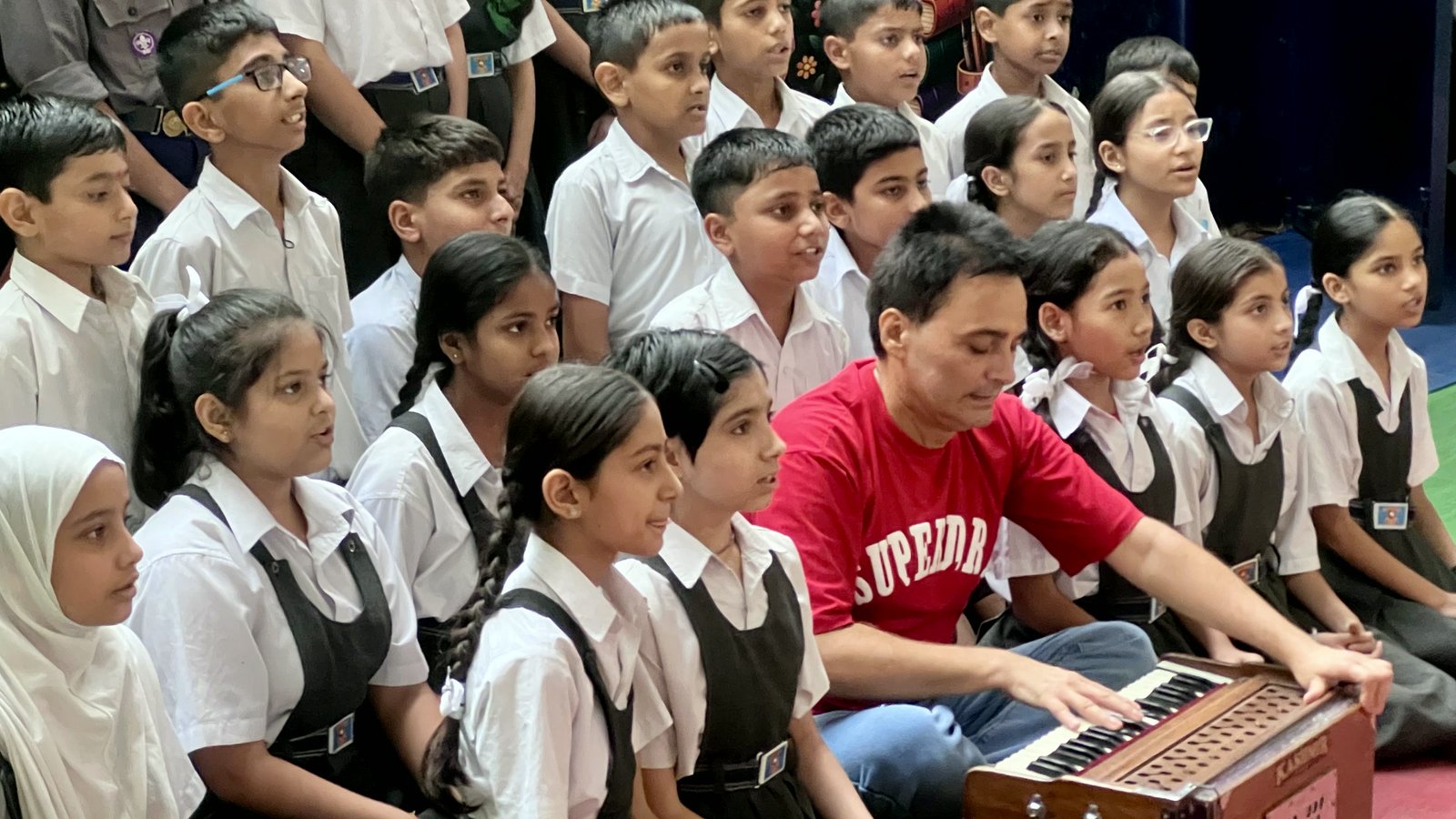 A teacher plays the harmonium while a class of boys and girls in school uniform sings around him