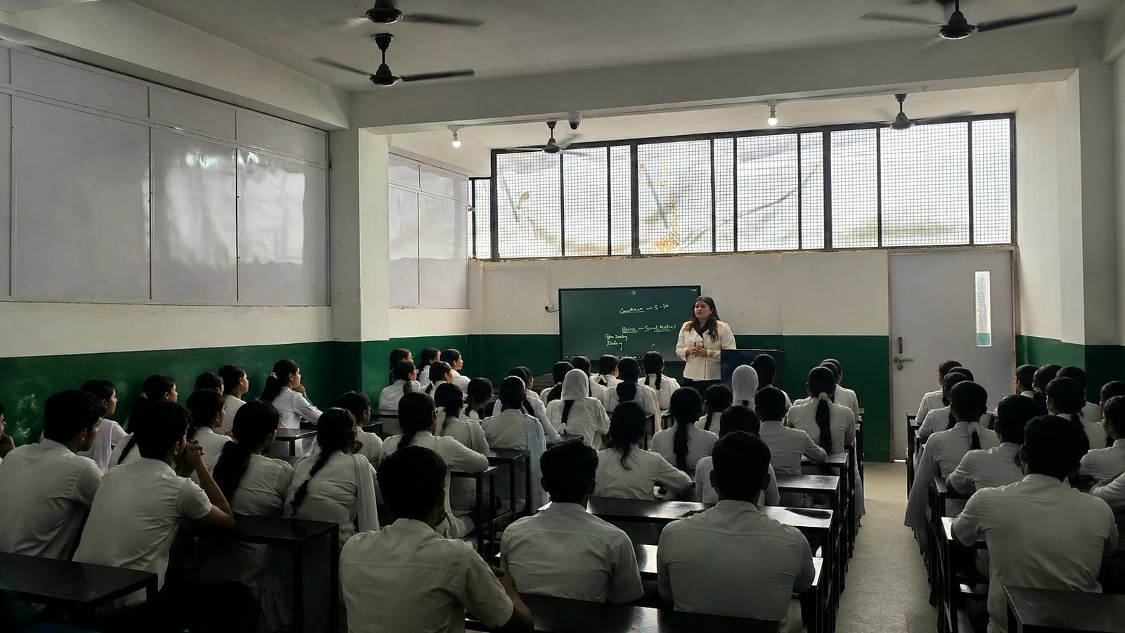 A full senior class at their desks facing the teacher, who is mid-lesson at the board at the front of the room