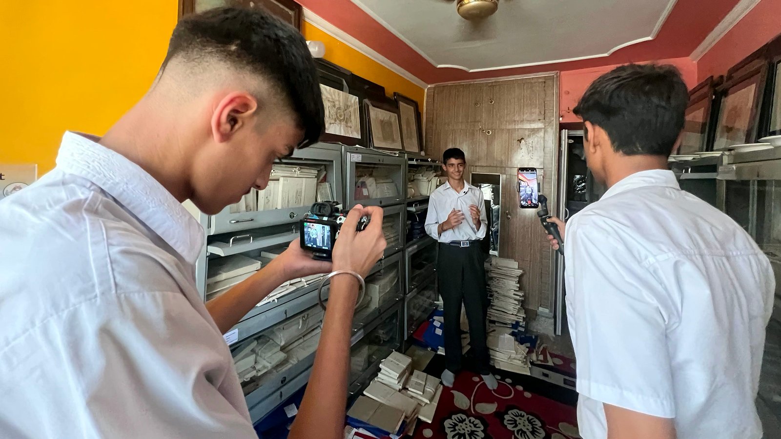 A senior student frames a shot on a camera while a classmate films on a phone gimbal and another student speaks to camera, among the school's record shelves