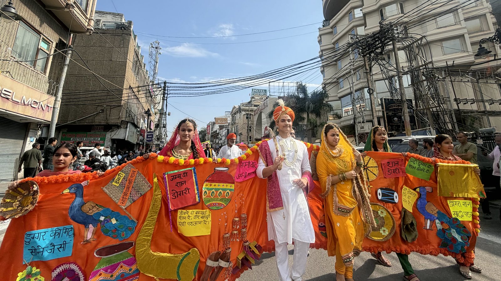 A student dressed as a king in white and a saffron turban walks through a Jammu market, his long orange robe of handmade Dogra symbols and Dogri placards held out by students in traditional dress