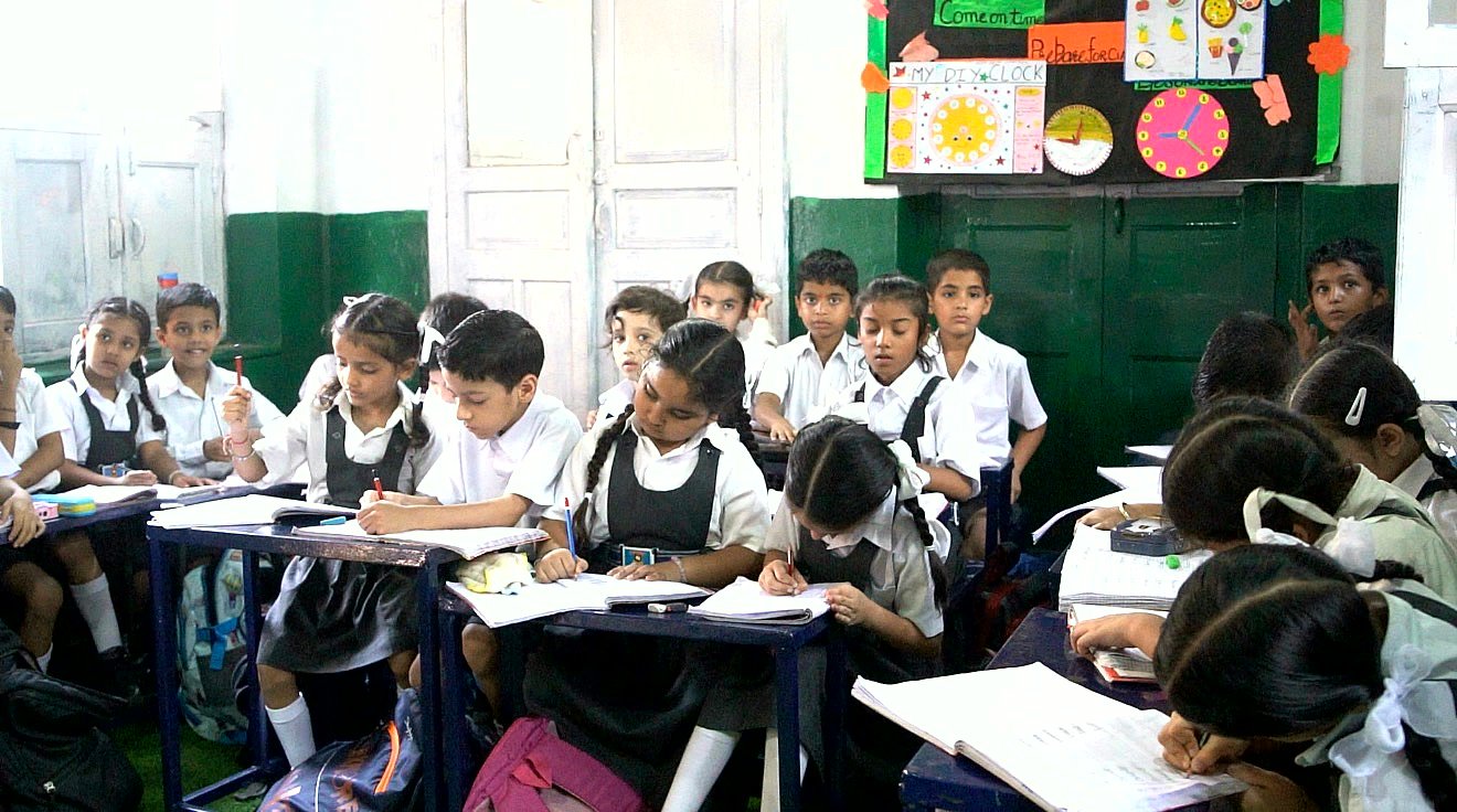 A full junior classroom in uniform working at their desks, a display board of pupil-made clocks on the wall behind them