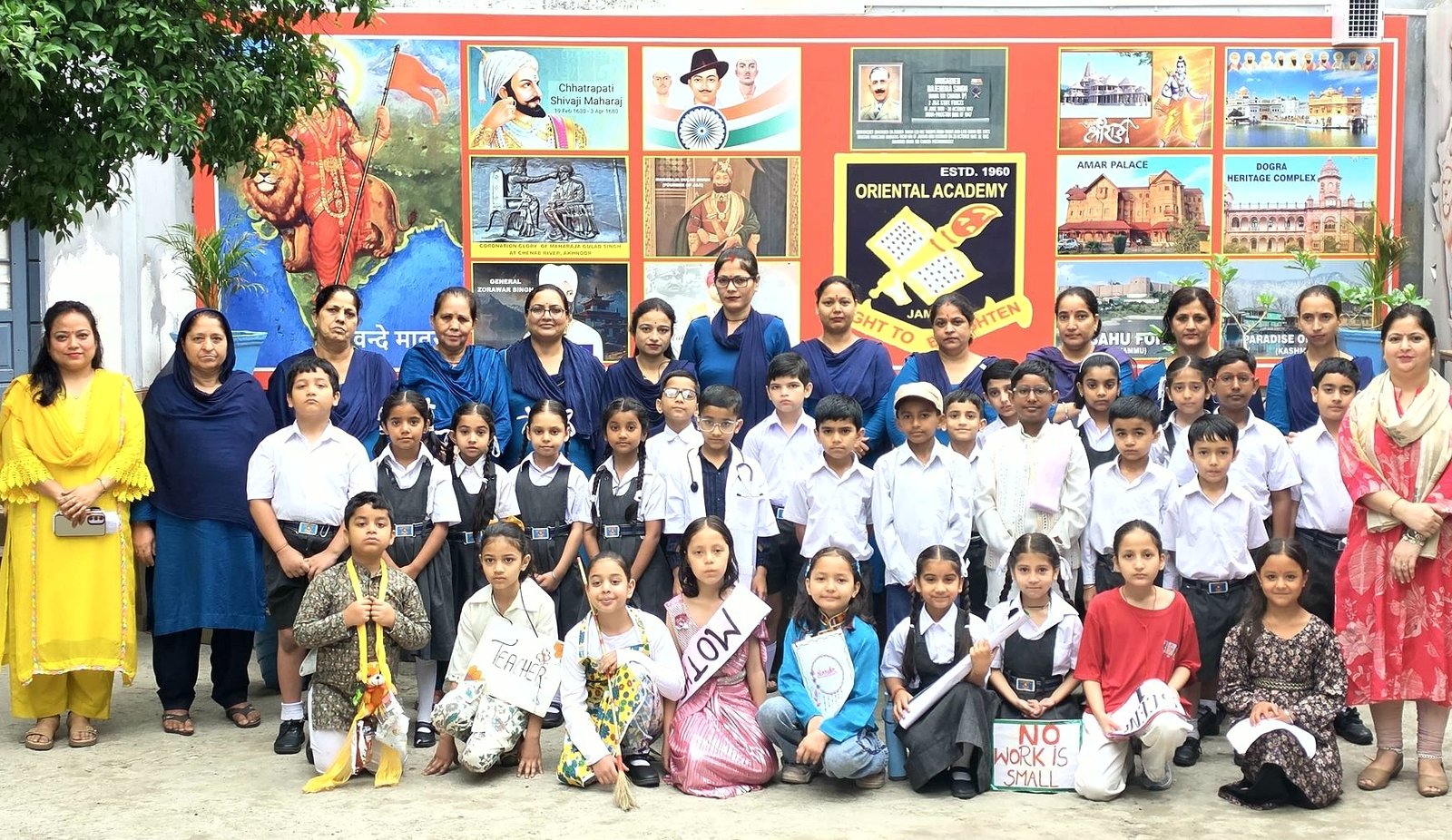 Junior-wing children dressed as community helpers, standing with their teachers in front of the school crest and a wall of Jammu&rsquo;s heritage