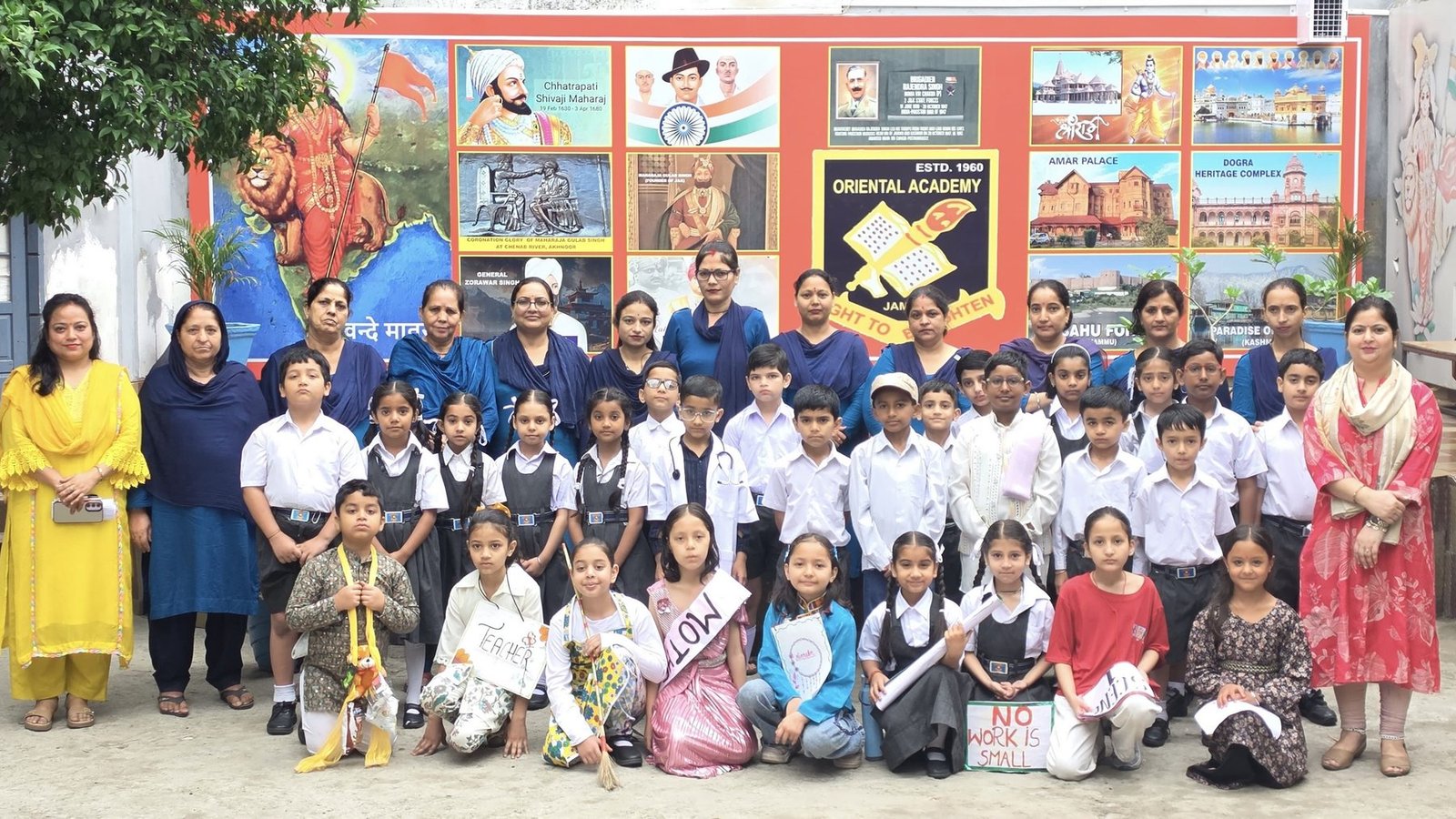 A junior class and their teachers in front of the school&rsquo;s painted heritage wall, the children dressed as the people who keep a town running, one holding a sign reading No work is small