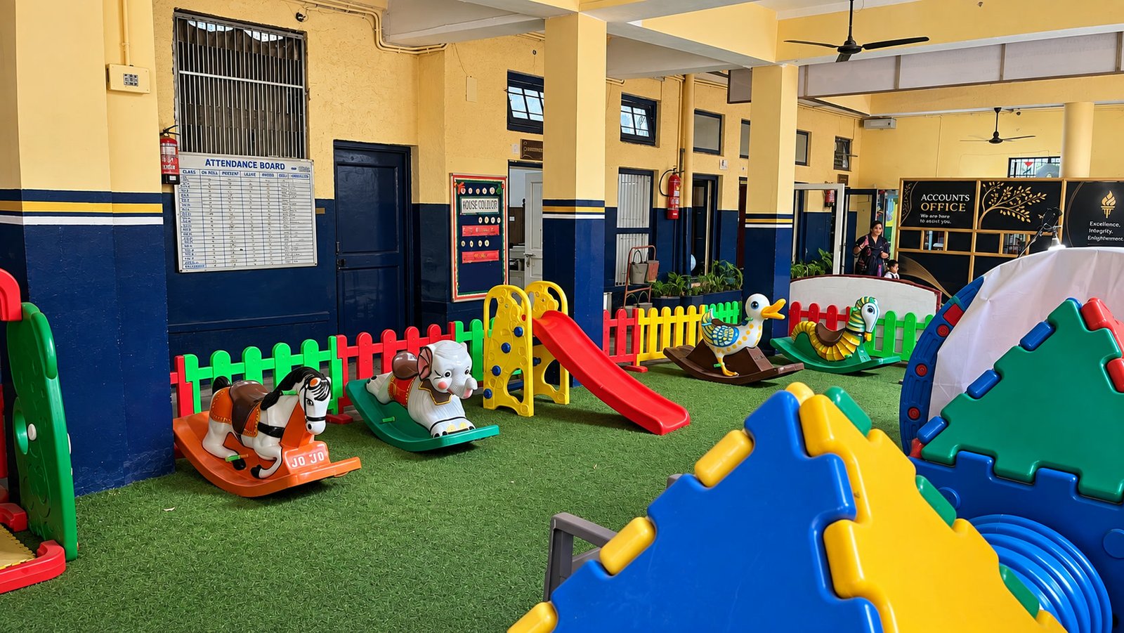 The junior play area on a green play floor: rocking horses, a slide and climbing frames, with the attendance board and the accounts office behind