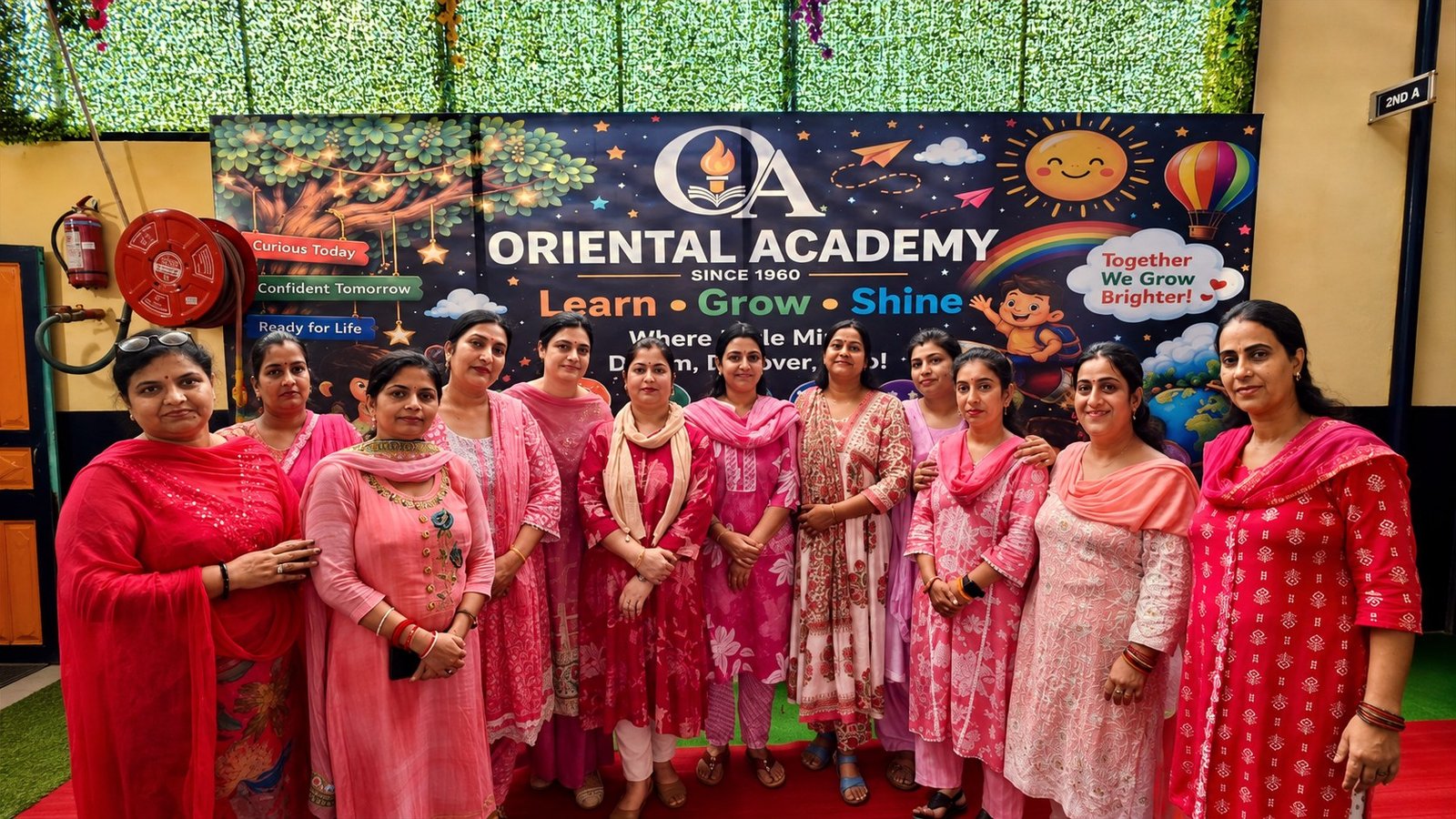 Thirteen junior-wing teachers standing together in shades of pink in front of the school banner