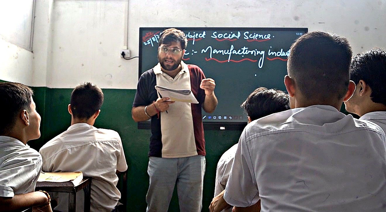 A teacher facing his class mid-sentence with his notes in one hand, the day&rsquo;s Social Science topic written up on the board behind him, a student turned towards him