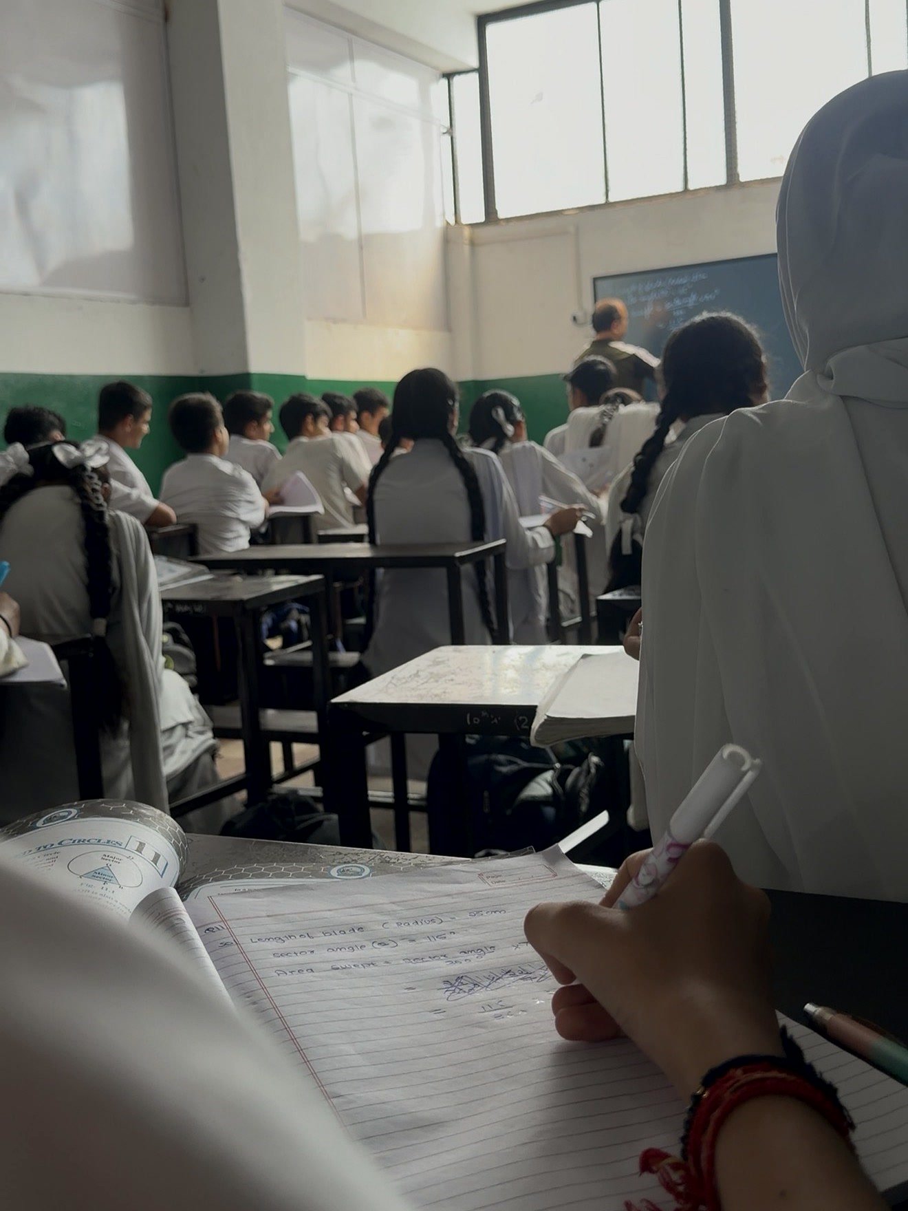 A maths lesson seen from a student&rsquo;s desk, a hand taking notes on circles in an exercise book, the class facing the teacher at the board