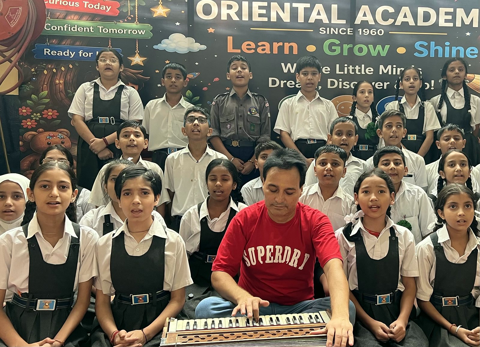 A class singing together behind their music teacher at the harmonium, the Oriental Academy banner behind them