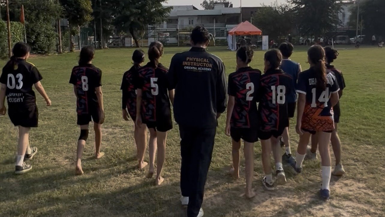 Oriental Academy girls in rugby kit walking out across the ground behind their physical instructor, whose tracksuit reads Physical Instructor, Oriental Academy
