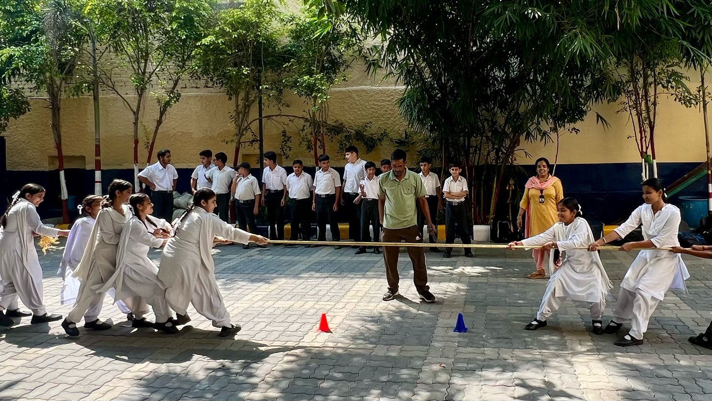 Girls in white salwar kameez pull hard on a tug-of-war rope while a teacher referees and a line of boys watches