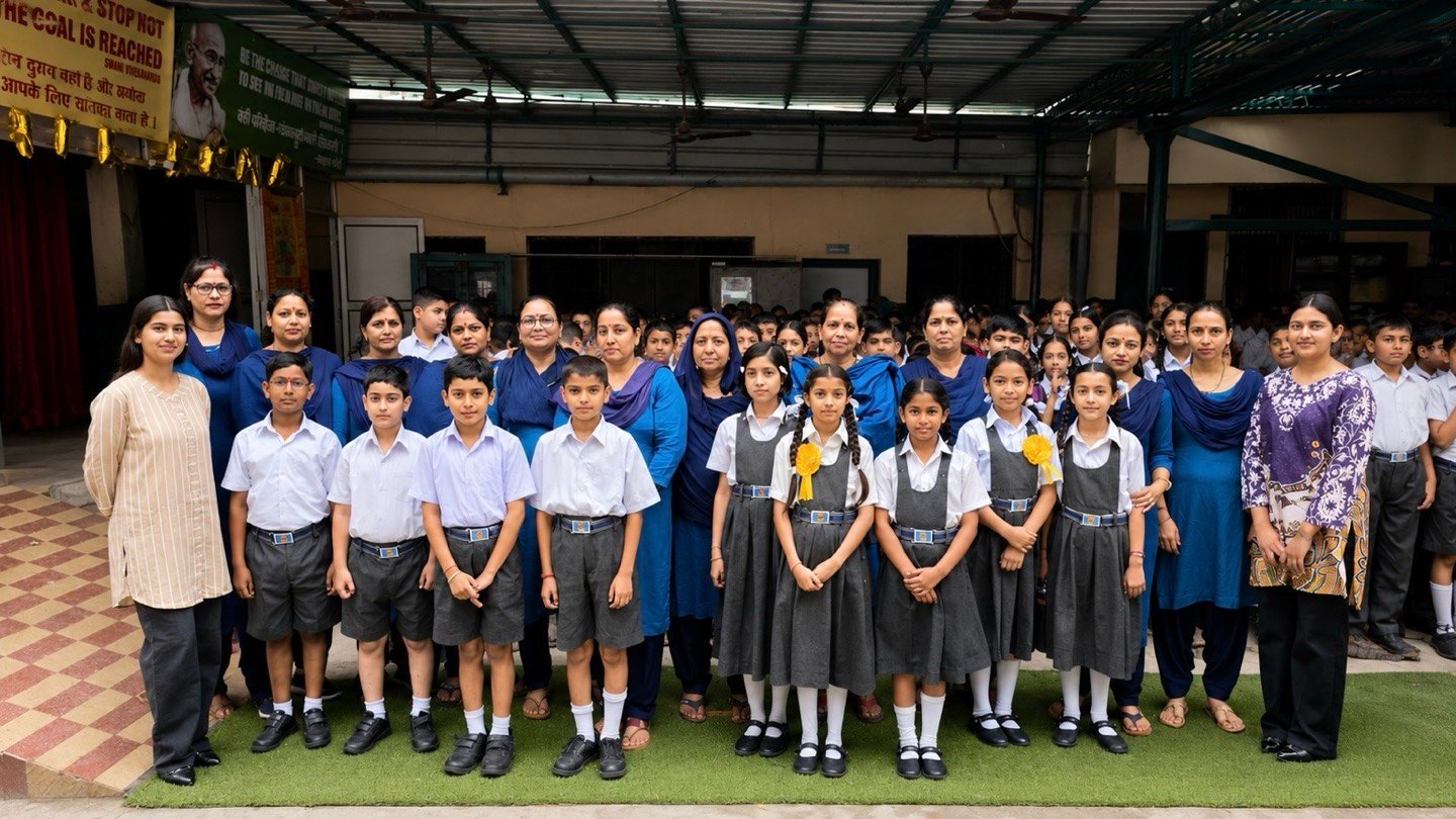 A line of students in today's uniform standing in front of their teachers, more students gathered behind them under the covered roof