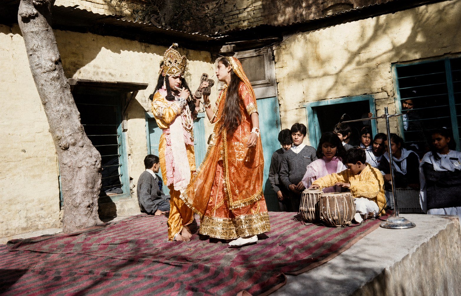 Two children in costume performing on a raised mud platform in the school yard in the 1960s, classmates seated on the ground around them